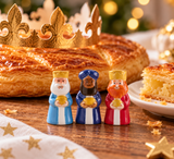 Trois fèves Rois Mages de la Nativité en céramique de 4 cm, aux tenues bleues et rouge, devant une galette des rois dorée et sa couronne, pour l'Épiphanie.